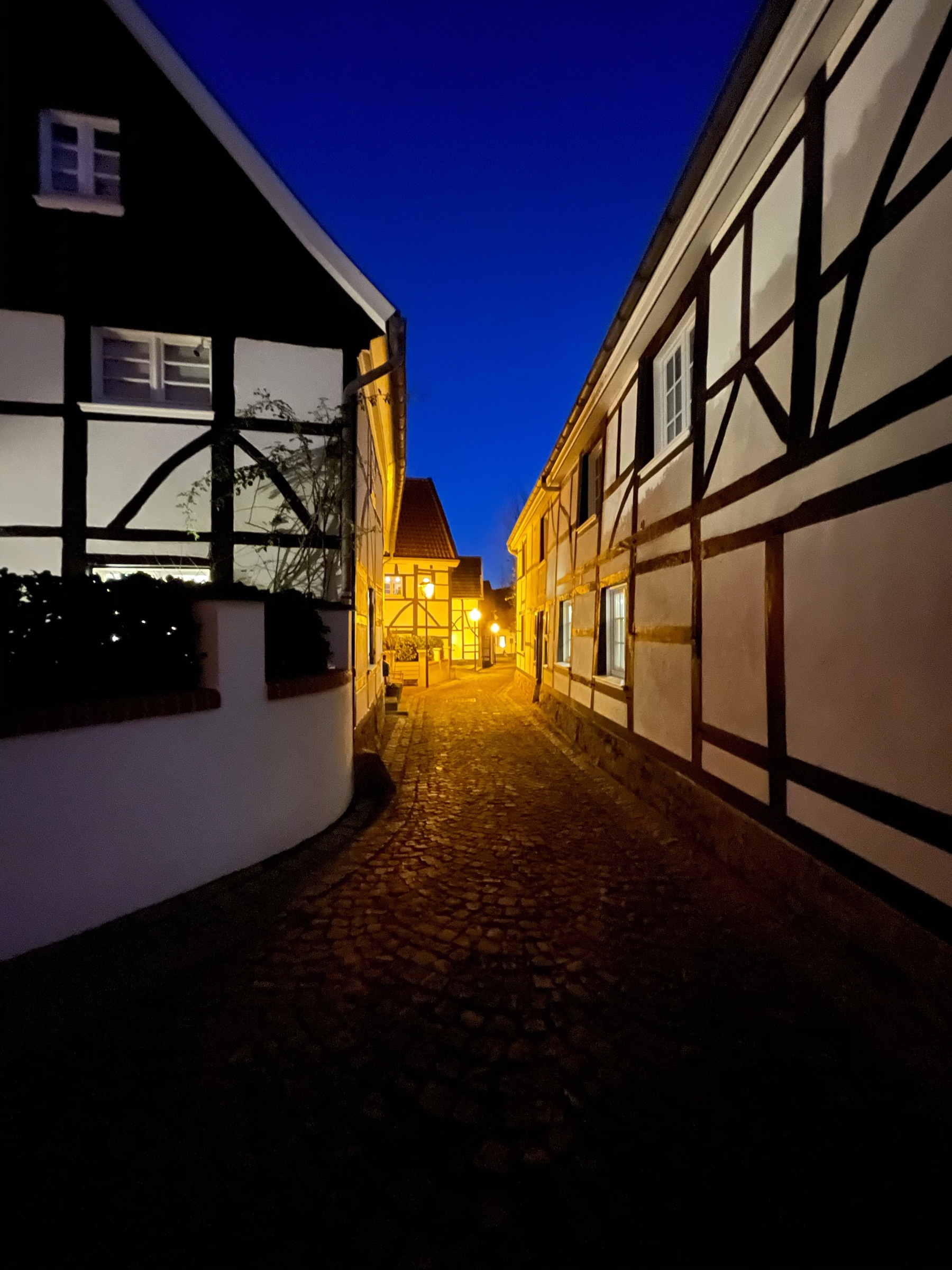 Narrow cobblestone street with framework houses on either side, shot at night with a dark blue sky and yellow lights in the distance. 