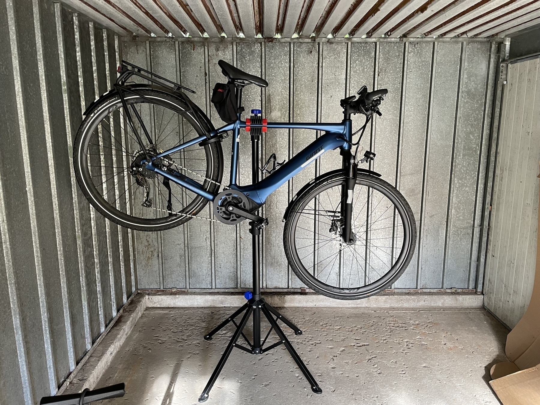 Blue electric bike suspended on a bike holder in mid-air. The bike holder is a black bar with four legs, on top is another bar at an 90 degree angle and a clamp holding the bike right over the motor for the center of gravity. The whole scene is inside a metal garage.