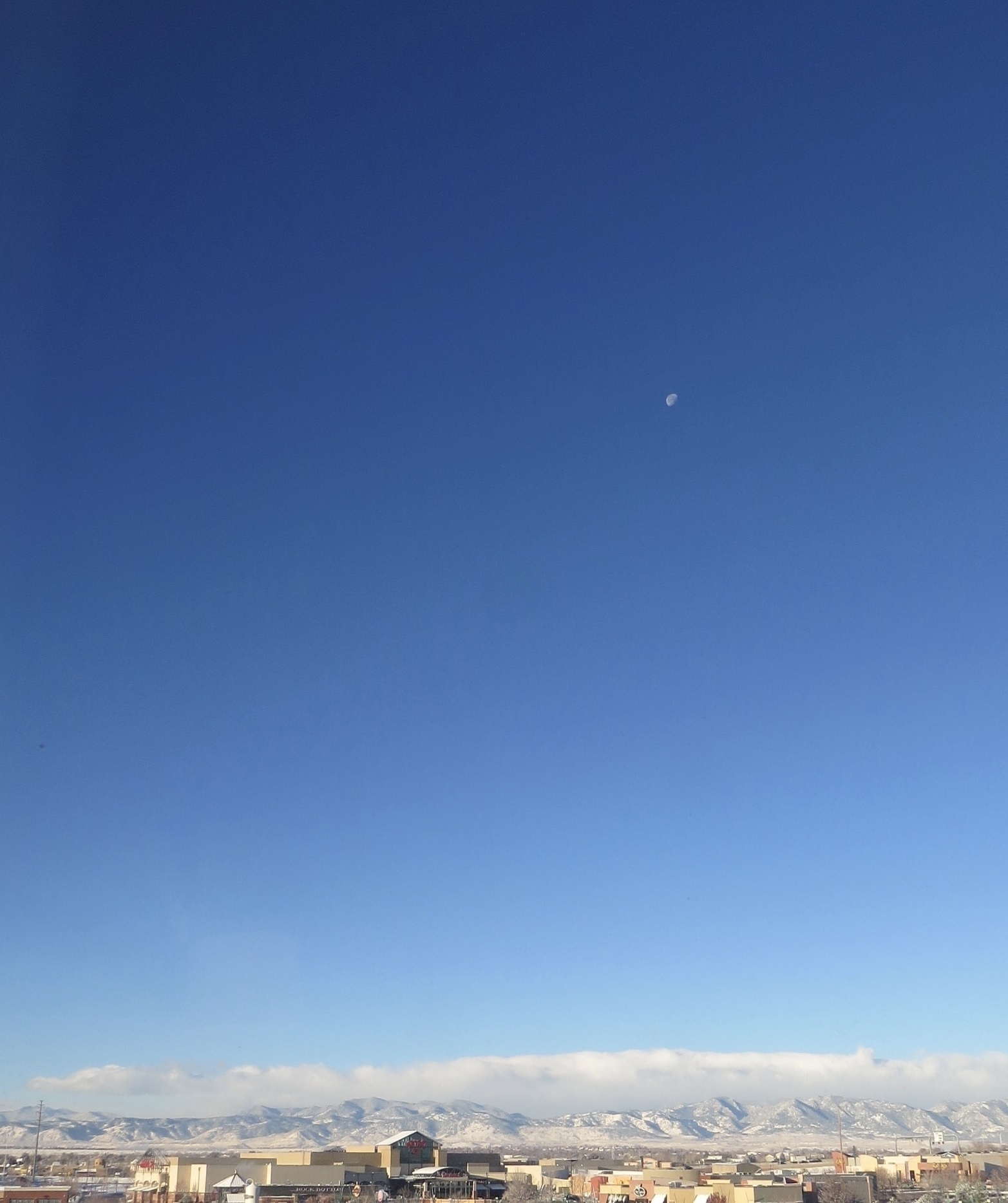 Over very low housing, the Rockies are seen on the bottom of the screen. Snow-covered caps. A cloud layer aboe them separating them from the crystal clear blue sky. In the sky a lone white dot, the moon, is seen on the top right of the image.